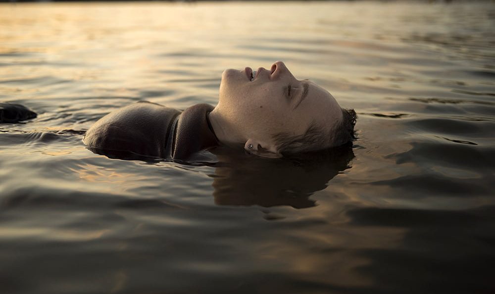 Shira Haas como Esty, flutuando em um lago em cena de Nada Ortodoxa