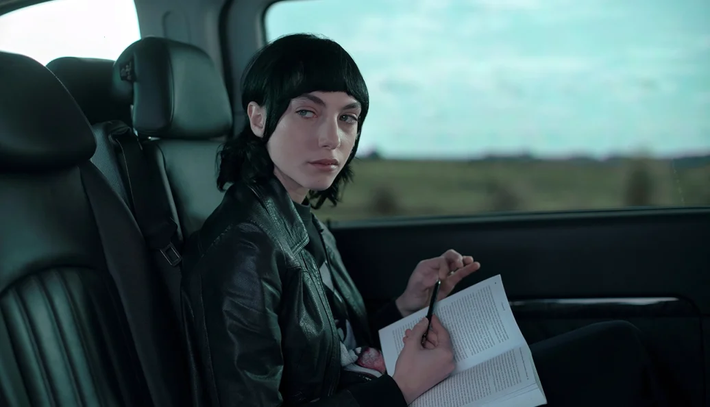 Cena de Irma Vep: mulher de jaqueta preta com um livro aberto no banco de um carro.