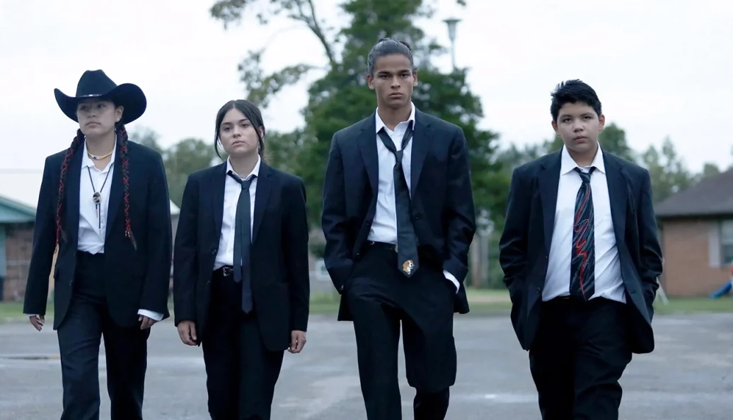 Cena de Reservation Dogs: quatro jovens de ternos escuros caminham lado a lado ao ar livre.