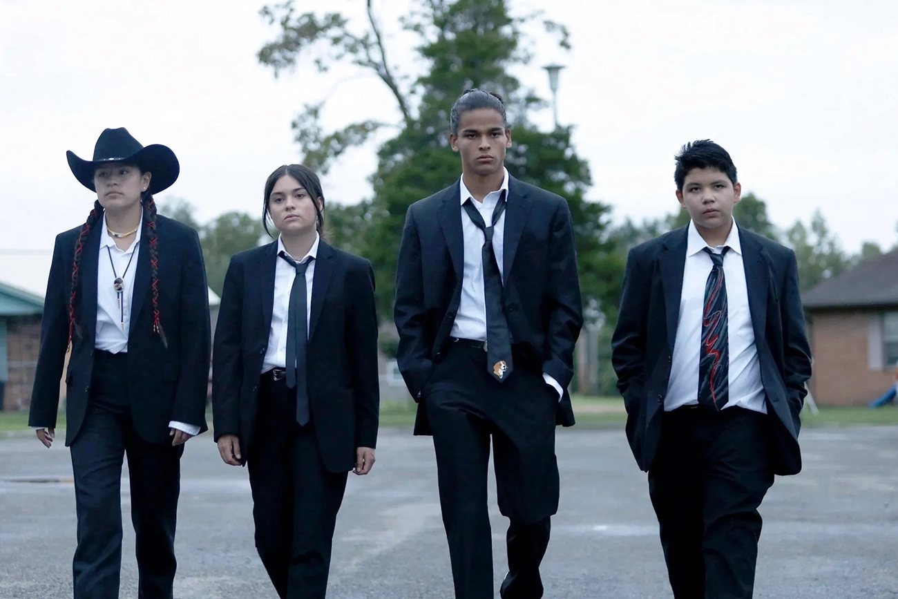 Cena de Reservation Dogs: quatro jovens de ternos escuros caminham lado a lado ao ar livre.