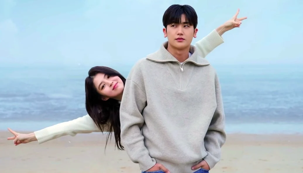 Mulher fazendo gestos com as mãos atrás de um homem em uma praia, em imagem de Soundtrack #1.