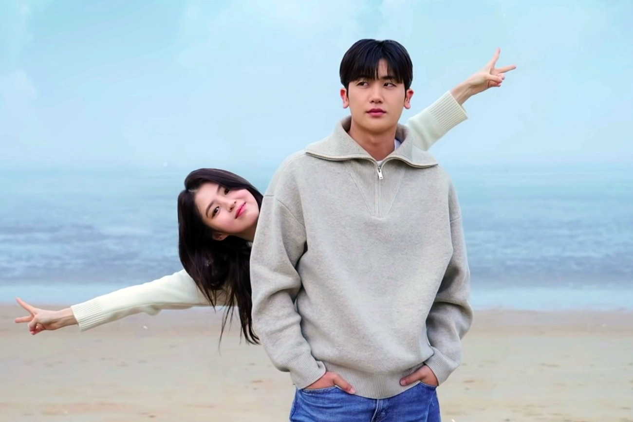 Mulher fazendo gestos com as mãos atrás de um homem em uma praia, em imagem de Soundtrack #1.