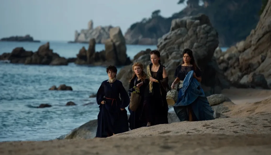 Quatro mulheres com vestidos de época caminham numa praia rochosa em cena de A Imperatriz.