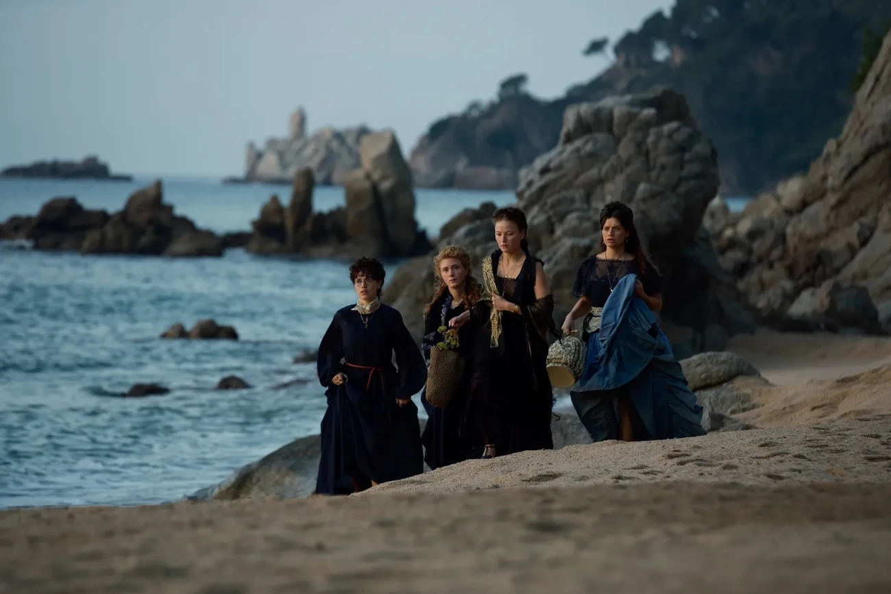 Quatro mulheres com vestidos de época caminham numa praia rochosa em cena de A Imperatriz.