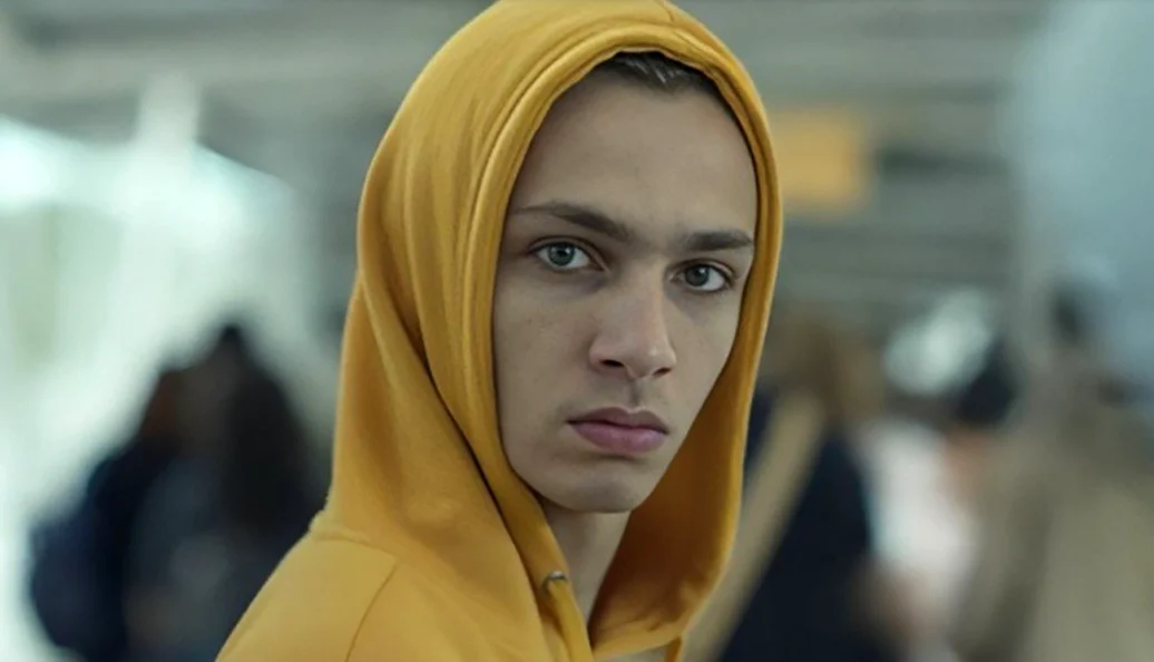 Personagem de Os Outros com capuz amarelo, em meio a uma multidão