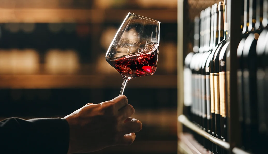 Mão segurando uma taça de vinho tinto diante de uma prateleira de garrafas.