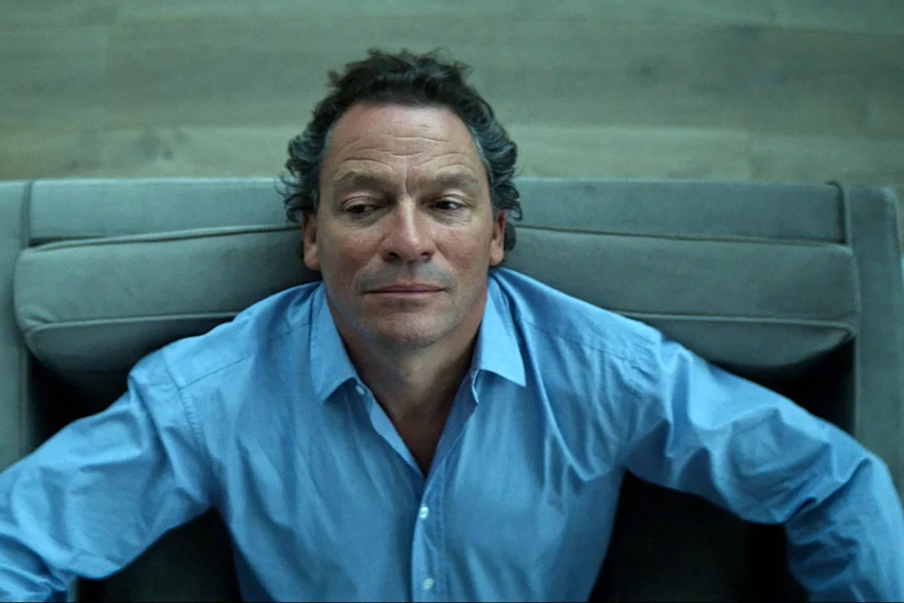 Dominic West sentado em uma poltrona.